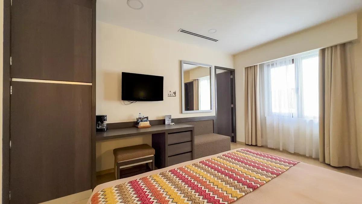 Wide view of the family room from Hotel Catedral. There is an entrance door to the other room with two beds. The colors are warm. Wooden floors. TV and working desk.