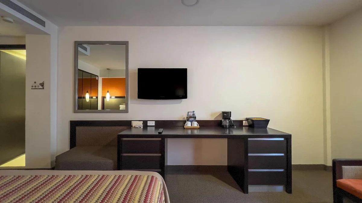 Quarto Suíte Junior do Hotel Catedral. TV na parede e uma cadeira no quadro, além de uma mesa de trabalho e um espelho.